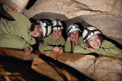 Four children in helmets with headlamps smile while exploring a cave, surrounded by rocky formations.