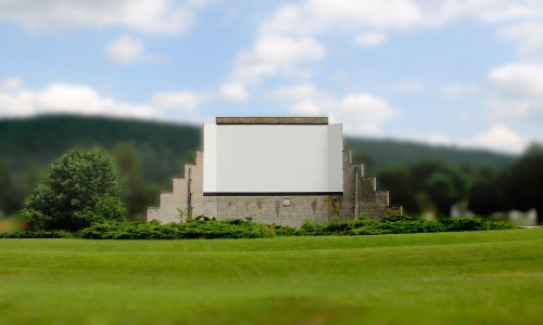A large, blank movie screen stands on a grassy field, surrounded by trees and hills in the background.