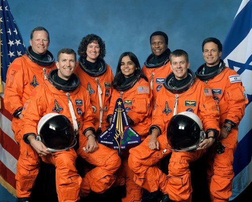 A group of seven astronauts in orange space suits, posing with flags and a mission patch, smiling against a blue backdrop.