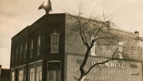 A vintage photo of a building with a bat silhouette on the roof and a bare tree in front.