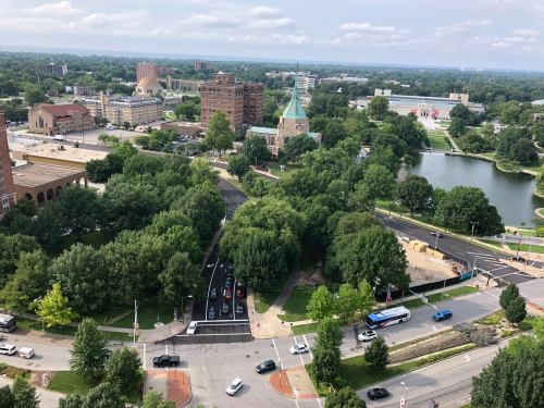 Aerial view of a cityscape featuring green trees, a pond, and buildings under a cloudy sky.