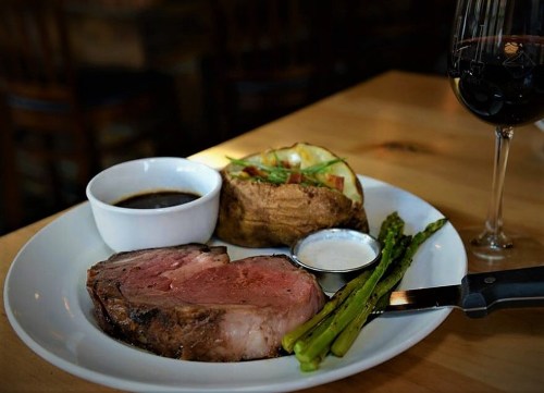 A plate with prime rib, baked potato, asparagus, dipping sauce, and a glass of red wine on a wooden table.