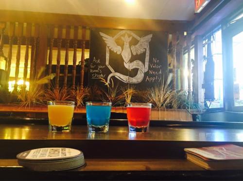 Three colorful shots (yellow, blue, red) on a bar counter with a chalkboard sign in the background.