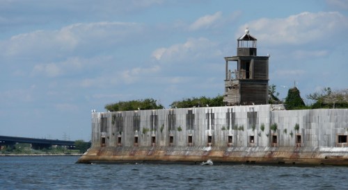 A weathered lighthouse on a stone pier, surrounded by water and greenery, under a blue sky with scattered clouds.