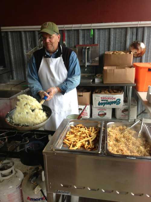 A man in an apron prepares food in a kitchen, with fries and a large pot of mashed potatoes on display.