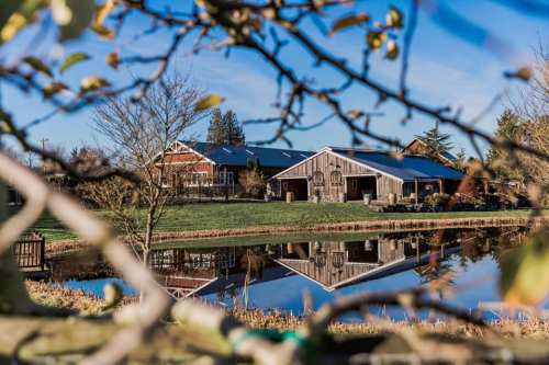 A rustic barn reflected in a calm pond, surrounded by trees and clear blue skies.