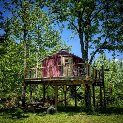 A purple yurt on stilts surrounded by lush green trees, with a deck and outdoor seating.