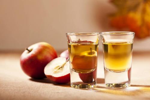 Two shot glasses filled with golden liquid, alongside whole and sliced red apples on a wooden surface.