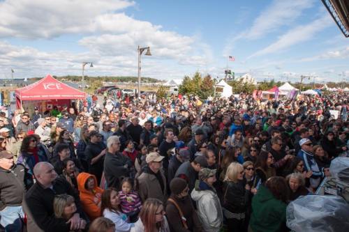 A large crowd gathers outdoors at a festival, with tents and a clear blue sky in the background.