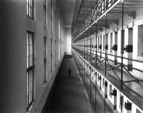 A black and white photo of a long, empty corridor in a prison, with tall windows and a solitary figure at the end.