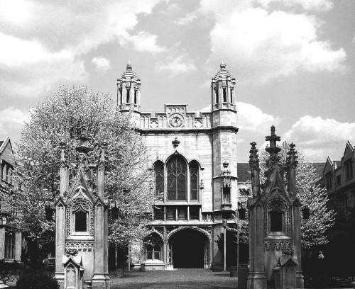 Historic building with ornate architecture, flanked by decorative gates and surrounded by blooming trees under a cloudy sky.