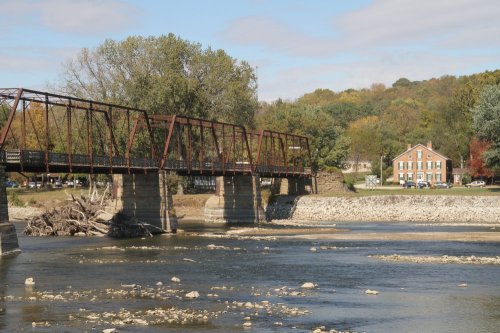 A rusted metal bridge spans a river, with trees and a red brick building visible on the shore in the background.