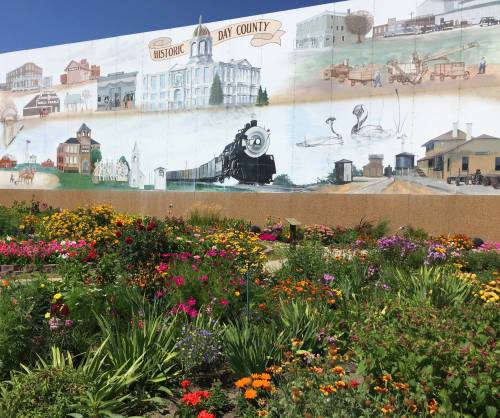 A colorful mural depicting historic scenes of Day County, surrounded by vibrant flowers in a garden.