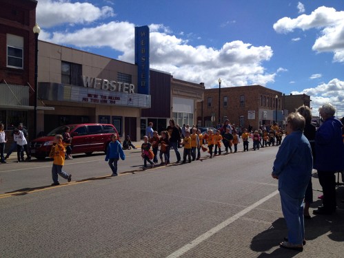 A parade with children in yellow shirts marching down a street, with spectators watching from the sidewalk.
