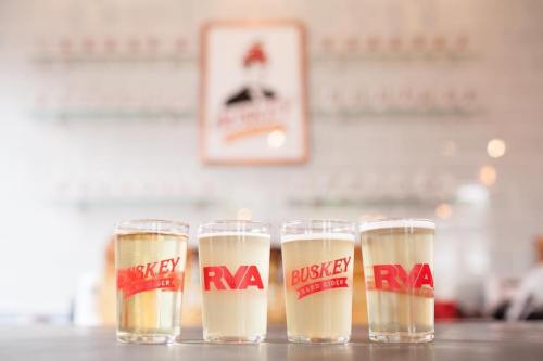 Four glasses of cider with "Buskey RVA" branding, displayed on a table with a blurred background.