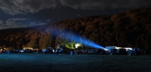 A nighttime scene of a parking area with cars, illuminated by a beam of light, surrounded by trees and a starry sky.