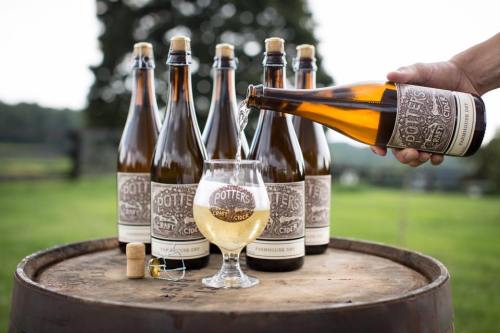 A hand pours cider from a bottle into a glass, surrounded by bottles on a wooden barrel in a scenic outdoor setting.