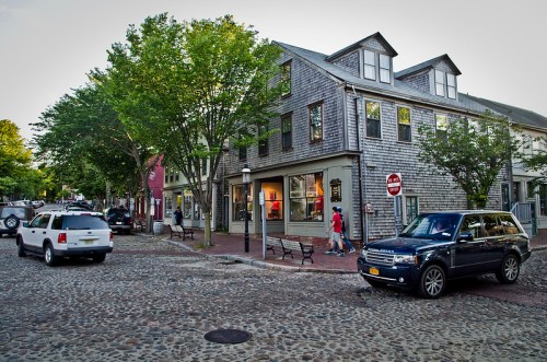 A quaint street scene with cobblestone pavement, shops, trees, and parked cars in a charming town.
