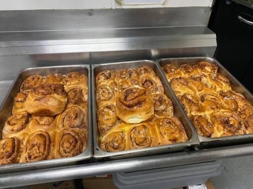 Trays of freshly baked cinnamon rolls, golden brown and swirled with cinnamon, cooling on a metal countertop.