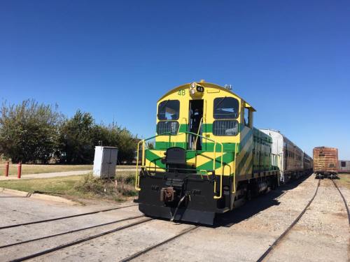 A green and yellow train engine parked on tracks under a clear blue sky, with trees in the background.