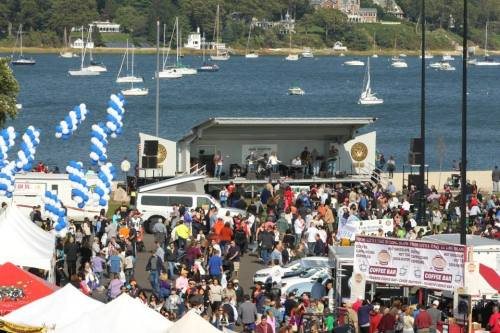 A lively outdoor festival with a crowd, food stalls, and a band performing on stage by a waterfront with boats.