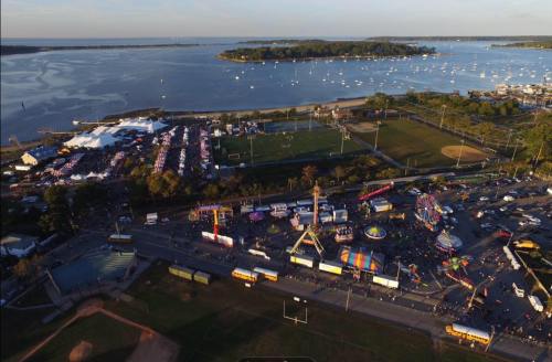 Aerial view of a waterfront fair with rides, tents, and a sports field, surrounded by water and boats in the distance.