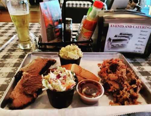 A tray with barbecue brisket, pulled pork, coleslaw, potato salad, a slice of bread, and a beer in the background.