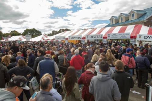 A large crowd gathers under striped tents at an outdoor festival, with signs for oysters visible.