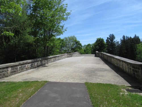 A wide, empty stone bridge surrounded by lush green trees under a clear blue sky.
