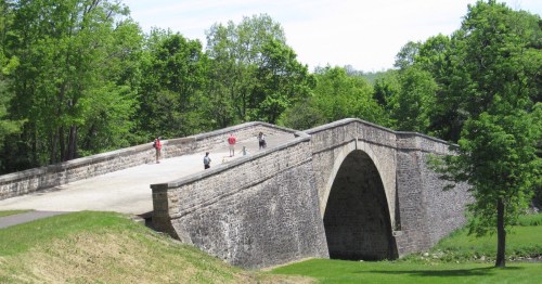 A stone arch bridge surrounded by greenery, with people walking on it and a grassy area in the foreground.