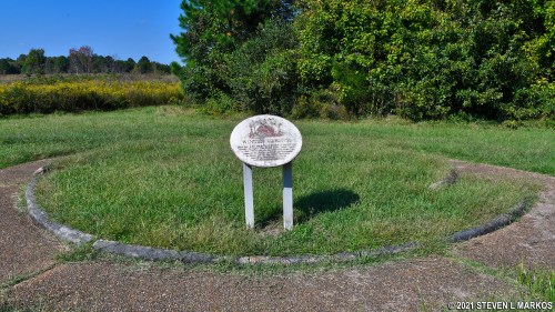 A circular stone marker in a grassy area, surrounded by trees and shrubs, with a plaque detailing historical information.