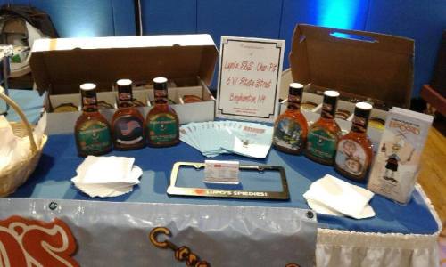 Display of various bottles of syrup on a table, with a sign and promotional materials in a blue setting.