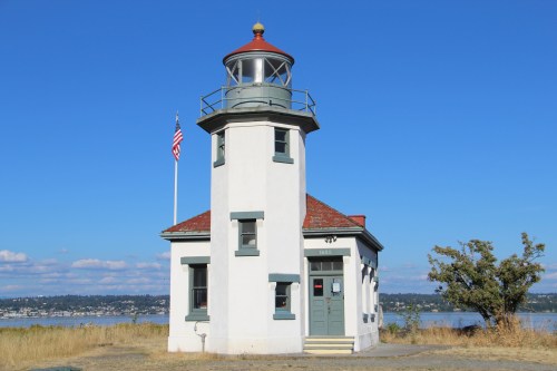 A white lighthouse with a red roof, surrounded by grass, against a clear blue sky and water in the background.