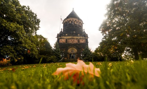A historic building with a dome, surrounded by trees and fallen leaves on a grassy foreground under a cloudy sky.