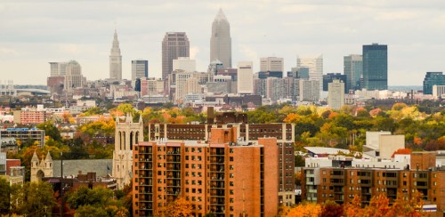 A panoramic view of Cleveland's skyline with autumn foliage in the foreground and a cloudy sky above.