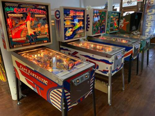 A row of vintage pinball machines, including "Cap Fantastic" and "Space Mission," in a retro arcade setting.