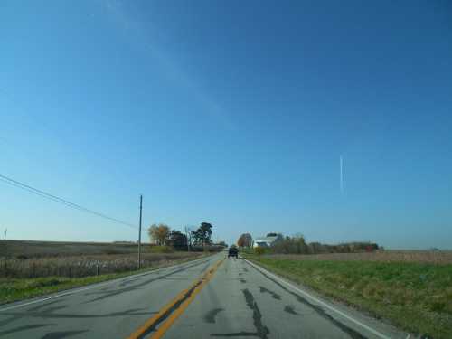 A straight rural road stretches into the distance under a clear blue sky, flanked by fields and a few trees.