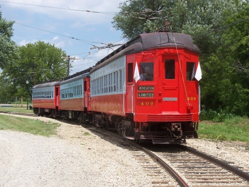 A vintage red train with blue accents on a track, surrounded by greenery on a sunny day.