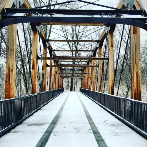 A snow-covered wooden bridge with metal supports, surrounded by bare trees in a winter landscape.
