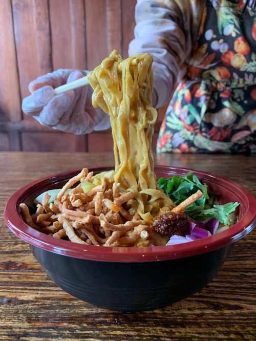 A hand holding chopsticks lifts noodles from a bowl filled with vegetables and crispy toppings.