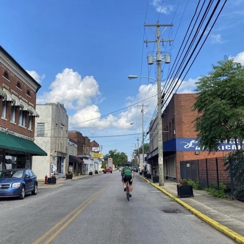 A cyclist rides down a quiet street lined with shops and power lines under a blue sky with fluffy clouds.