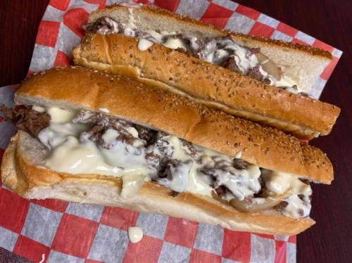 Two loaded sandwiches with sliced beef and creamy sauce on sesame seed hoagie rolls, resting on red and white checkered paper.