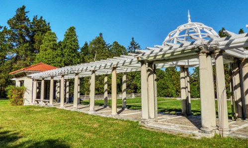 A white pergola with columns and a domed roof, surrounded by green grass and trees under a clear blue sky.