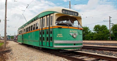 A vintage green and cream streetcar labeled "MUSEUM" parked on tracks, surrounded by greenery and a cloudy sky.