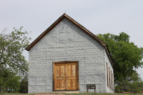 A rustic schoolhouse with wooden doors, set against a backdrop of trees and a cloudy sky. Sign reads "School House Open."