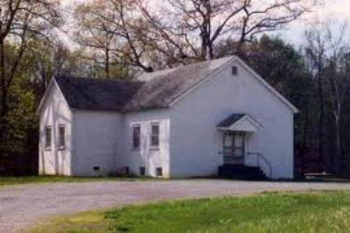 A small, white, single-story building with a sloped roof, surrounded by trees and grass.