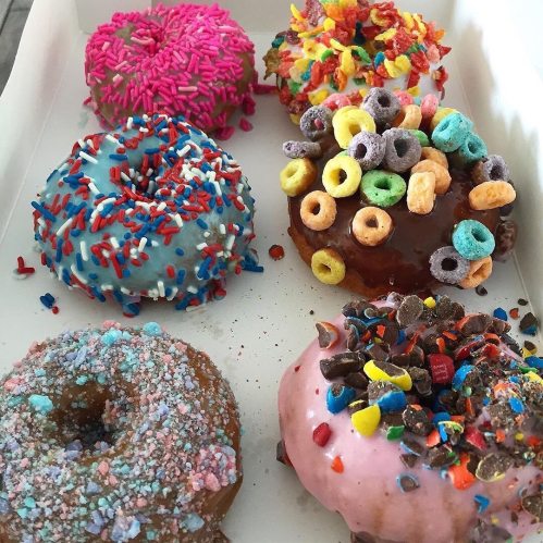 A box of colorful donuts topped with sprinkles, cereal, and chocolate bits in various flavors.