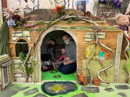 A family sits inside a whimsical, nature-themed playhouse decorated with plants and colorful elements.