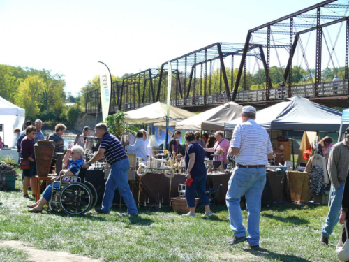 A bustling outdoor market with vendors and shoppers, set against a backdrop of a historic bridge.