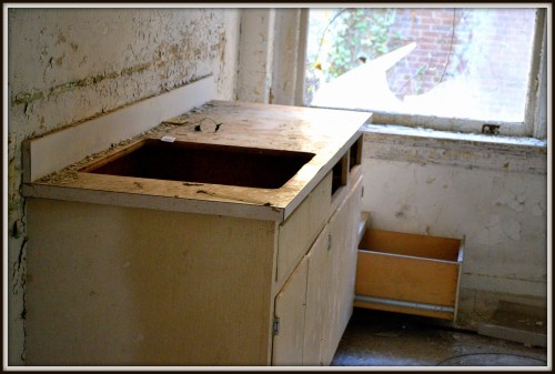 An old, dusty countertop with a sink, next to a window and a partially open drawer in a neglected room.